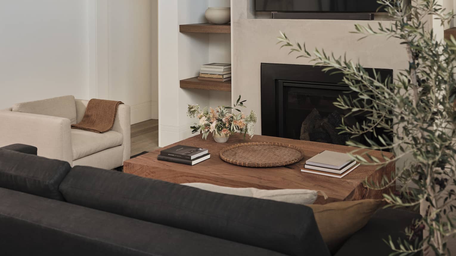 Cozy living room with beige armchair, dark gray sofa, wooden coffee table with books and flowers, built-in shelves with decor, fireplace and wall-mounted TV