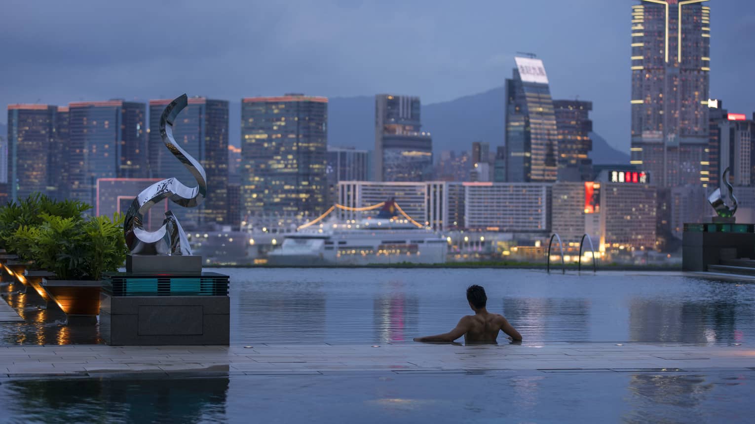 Person relaxing at the edge of an infinity pool overlooking a city skyline at dusk, with tall buildings and a cruise ship in the background.
