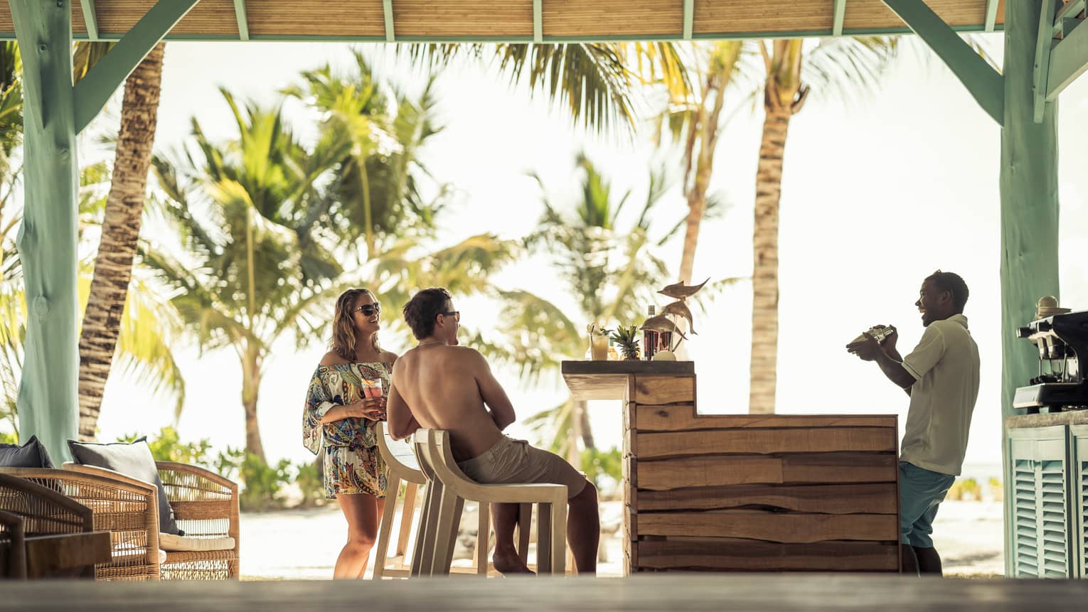 Couple in beach wear laugh with bartender who shakes cocktail in thatched roof bar