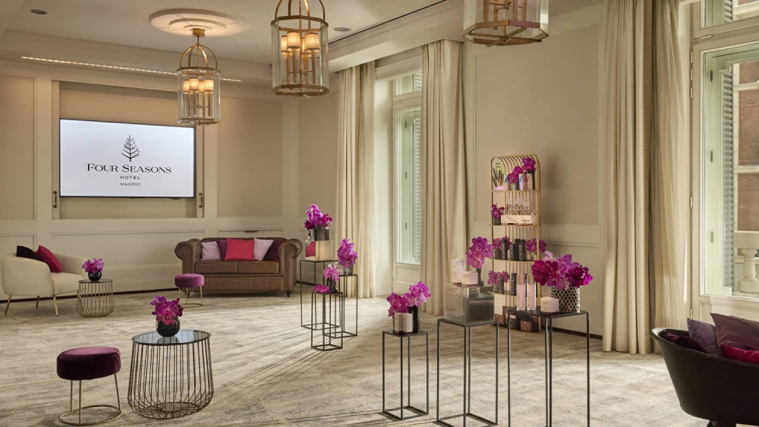 A meeting room with beige walls and carpet has large windows letting in natural light, chandelier lighting, a sofa and a chair, a flat-screen TV, plus a small a shelving unit and various small tables holding pink flowers in vases