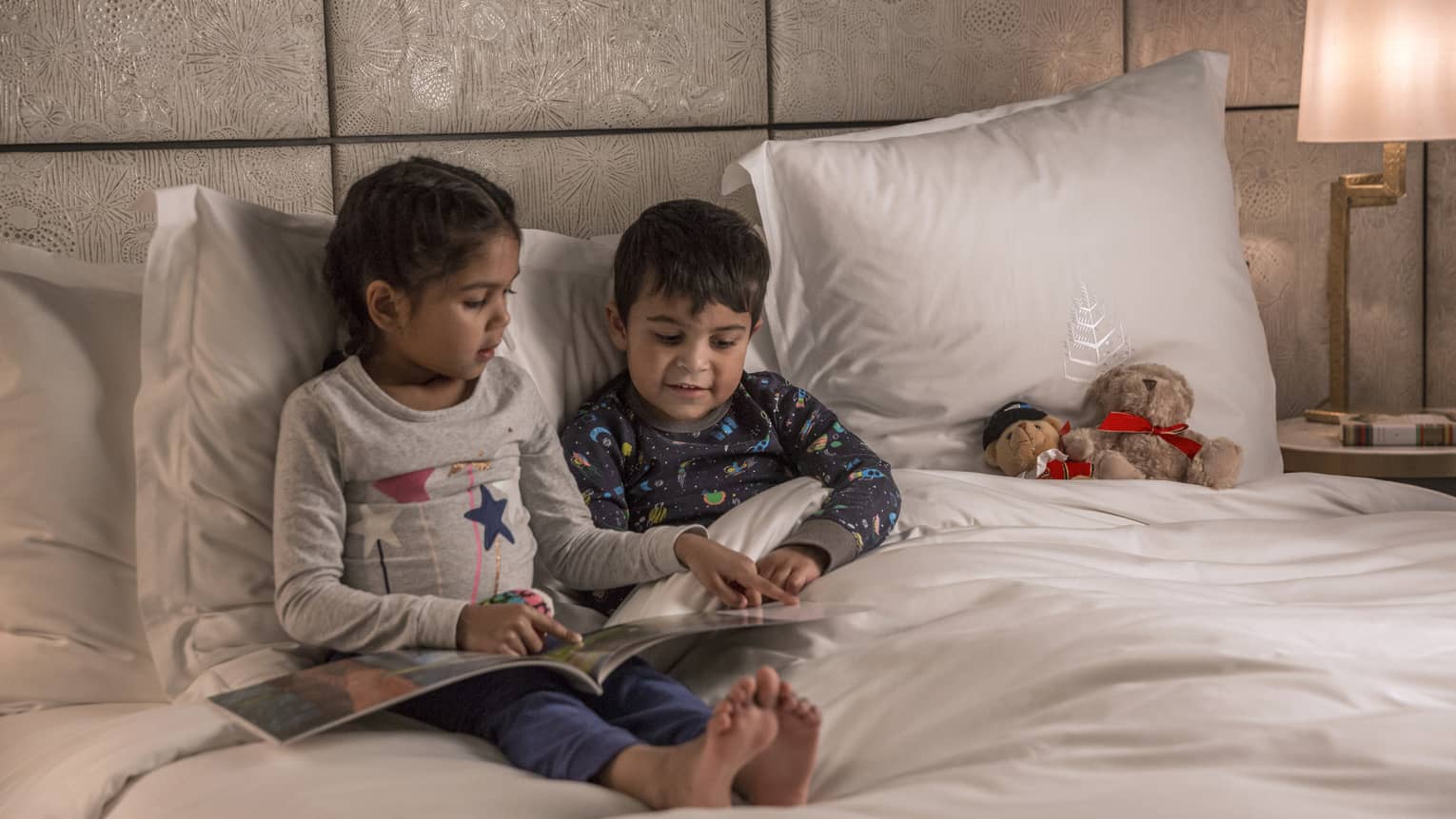 Young girl reads to her brother at bedtime, both in pajamas, a teddy bear propped on the pillow next to them