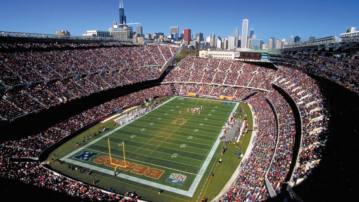 Aerial view of open stadium with football field, stands filled with fans against blue sky