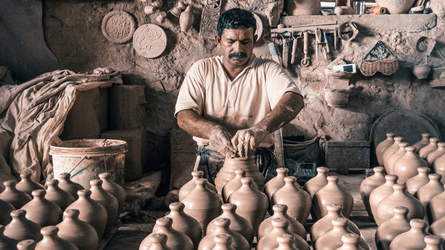 A man crafts pottery in a workshop with dozens of clay vases surrounding him.