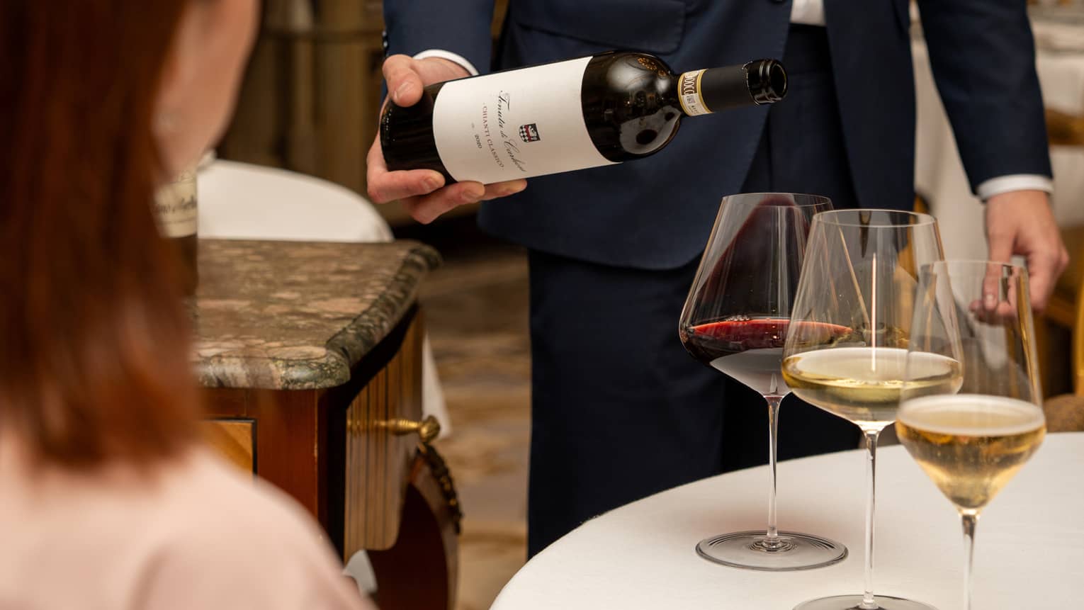 A guest sits at a table with three glasses of wine as a smiling sommelier tilts a bottle of red wine over one of the glasses.