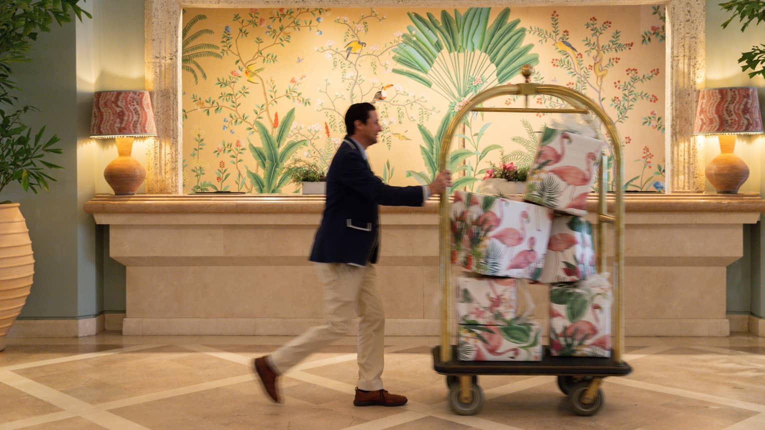 Bellhop wearing navy blazer and khaki pants pushes a luggage cart filled with presents through a hotel lobby