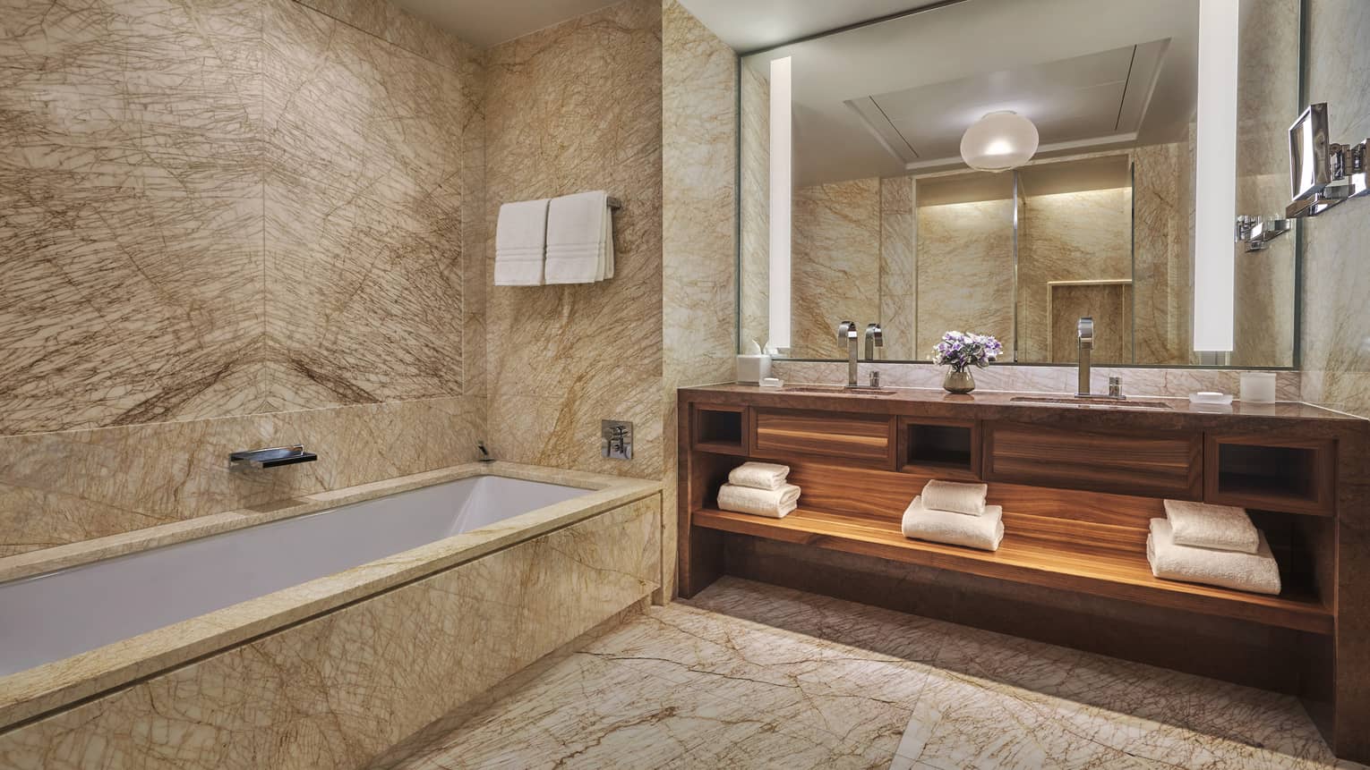 Luxury hotel bathroom with marble walls, a built-in bathtub, large mirror, double-sink wooden vanity with towels and soft lighting