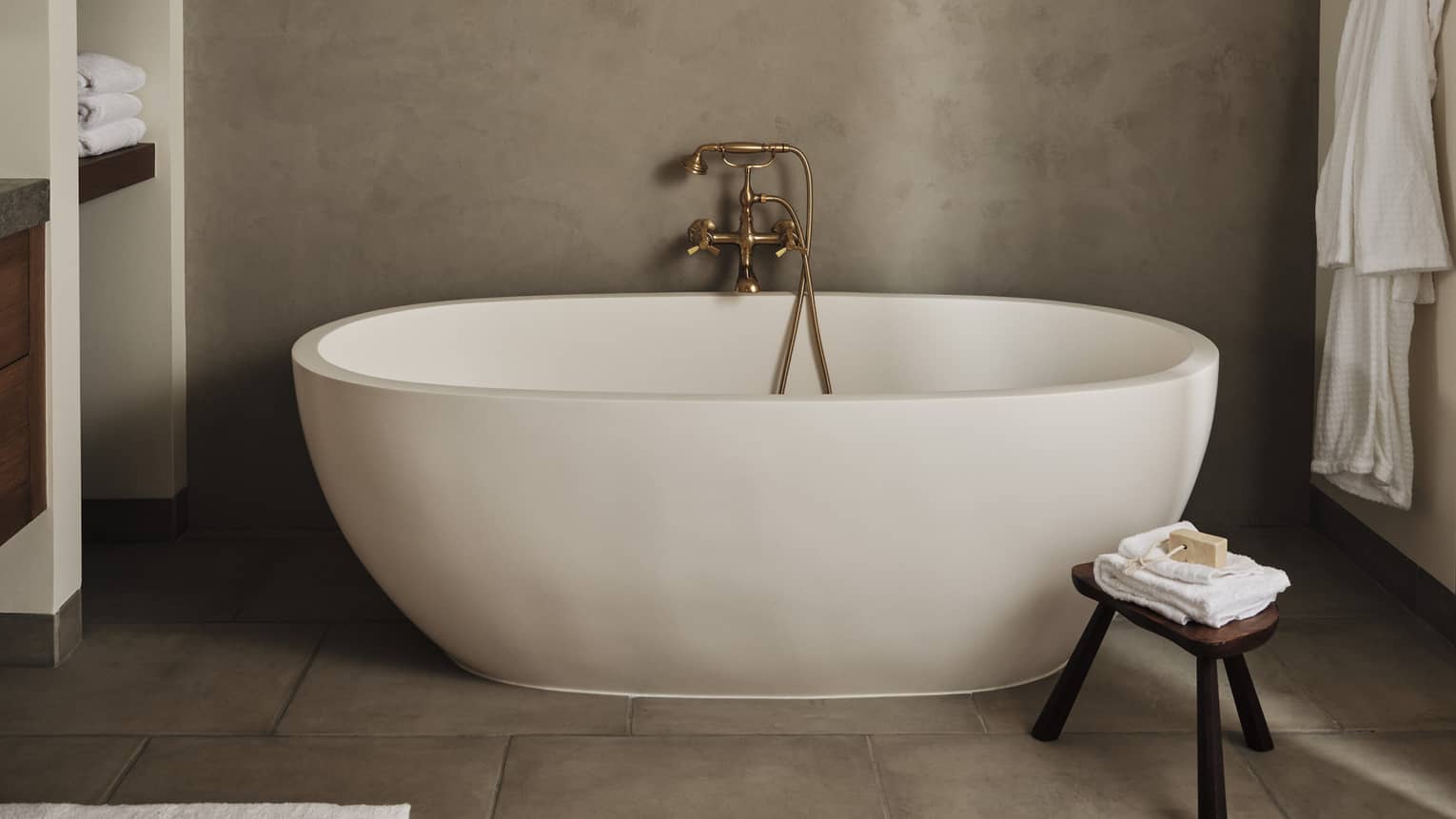 Luxury resort bathroom with a freestanding oval bathtub, built-in towel shelves, small wooden stool with folded towels and soap and bathrobe hanging on the wall