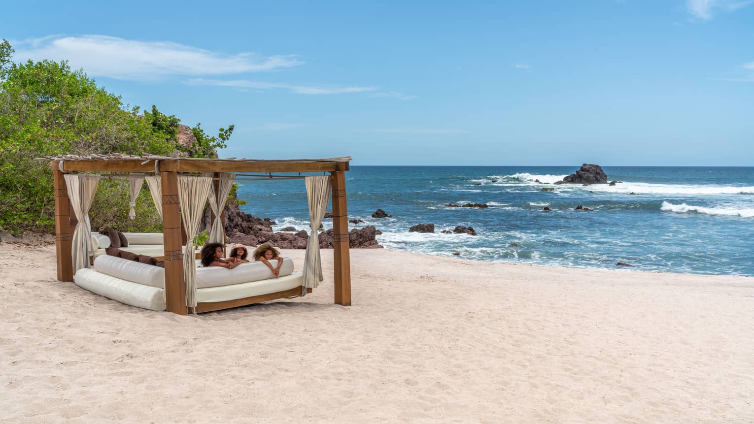 A cabana with three children laying on a plush seat underneath it on the beach, the ocean and plants are in the background.