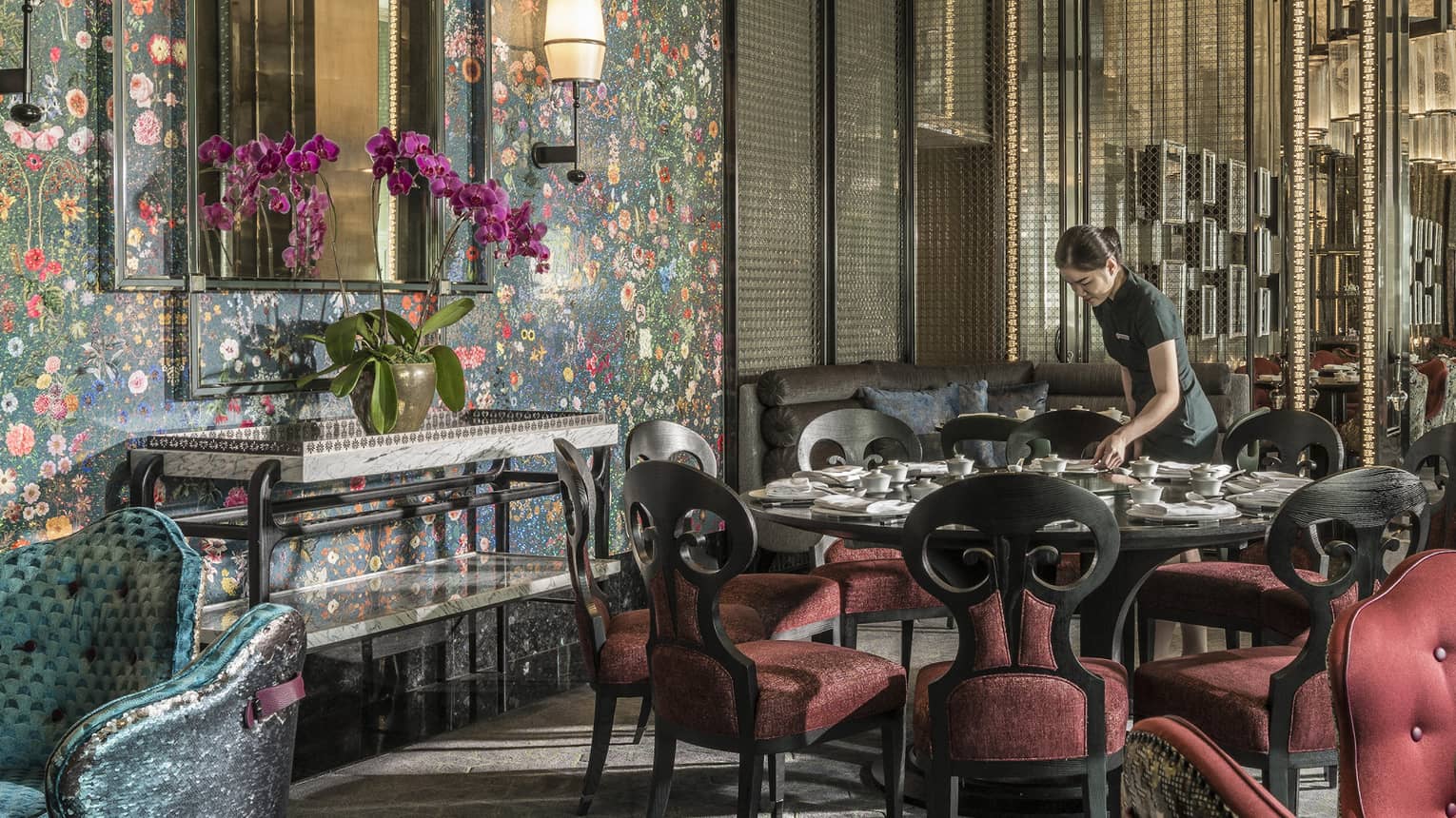 Hotel staff sets table with elegant red velvet chairs under tall mirror, floral wallpaper