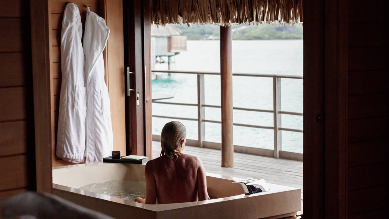 A guest relaxes in a large tub and looks past sliding wooden doors at the ocean. The water is lively and lit by the sun.