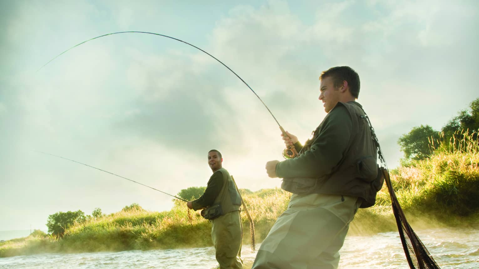 Two people fly fishing in a river, standing knee-deep in the water, surrounded by mist and greenery.