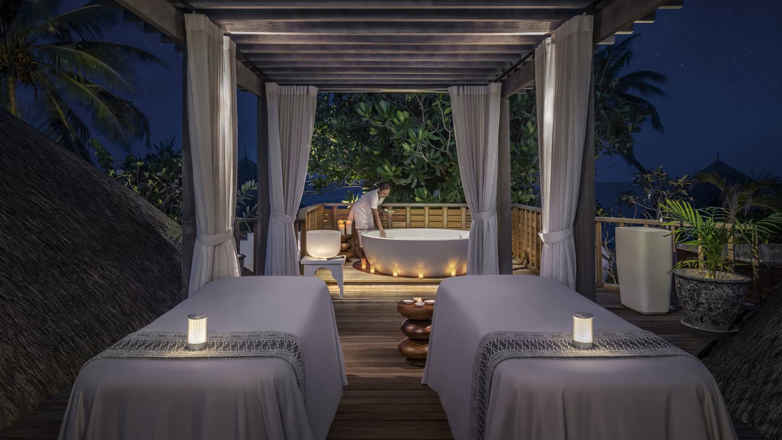 Outdoor night spa setting with two massage tables, soft candlelight, and a staff member preparing a bathtub under a canopy surrounded by tropical greenery