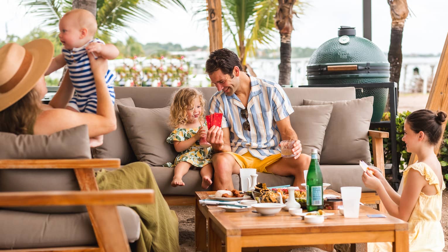 Family with two adults and three children ranging in age from infant to tween sit on grey outdoor sofas playing cards and enjoying food served on the coffee table in front of them