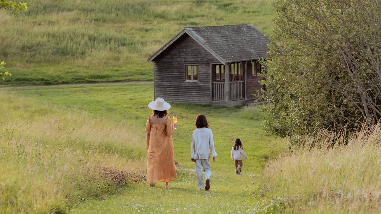 An adult and two children walk down a mown grass path in the middle of a field towards a wood cottage. Field grasses and scattered trees are in the background, along with a dramatic partly cloudy sky.
