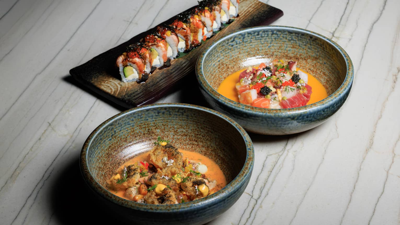Two stoneware bowls filled with food sit next to a rectangular platter of sushi on a white wood-grain table
