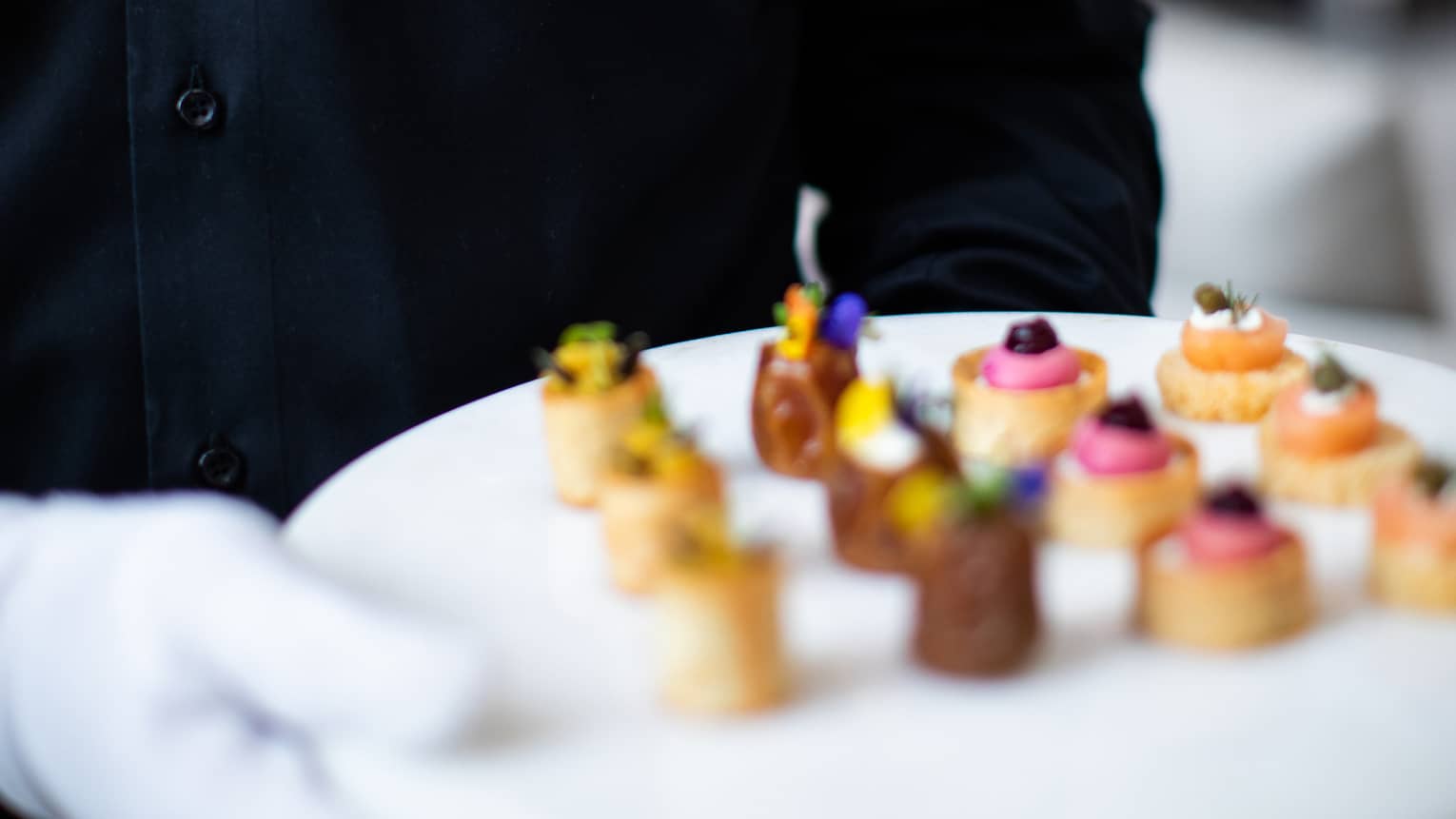 Waiter wearing white gloves holds a white round tray of small appetizers