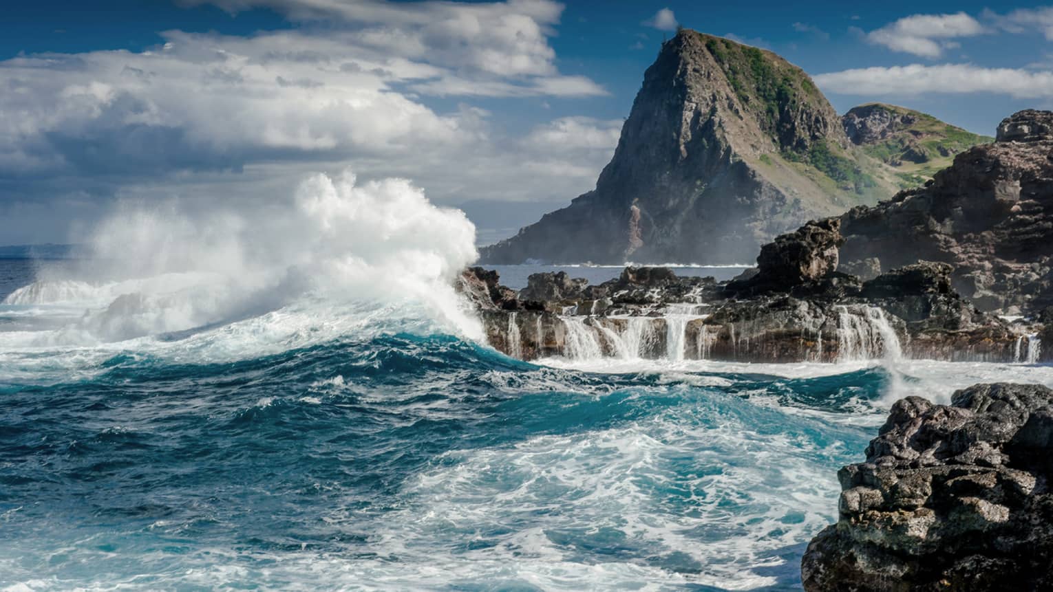 Crashing waves strike the shore of a volcanic island. A mountain looms in the background against a partially-cloudy sky.