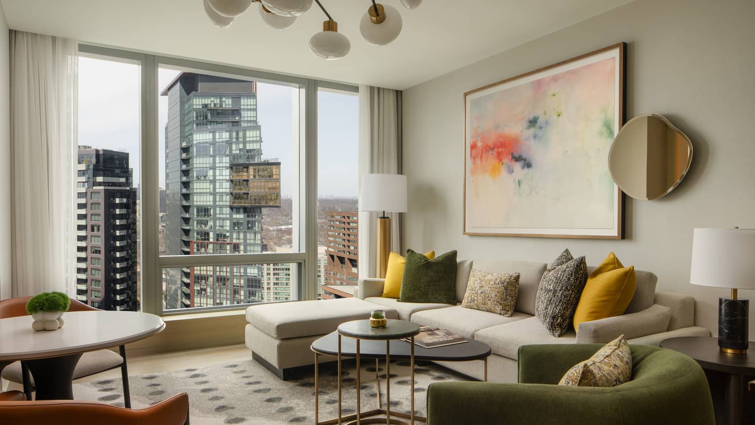 Stylish suite living area with a large sectional sofa, colorful pillows, modern artwork, and floor-to-ceiling windows overlooking the city