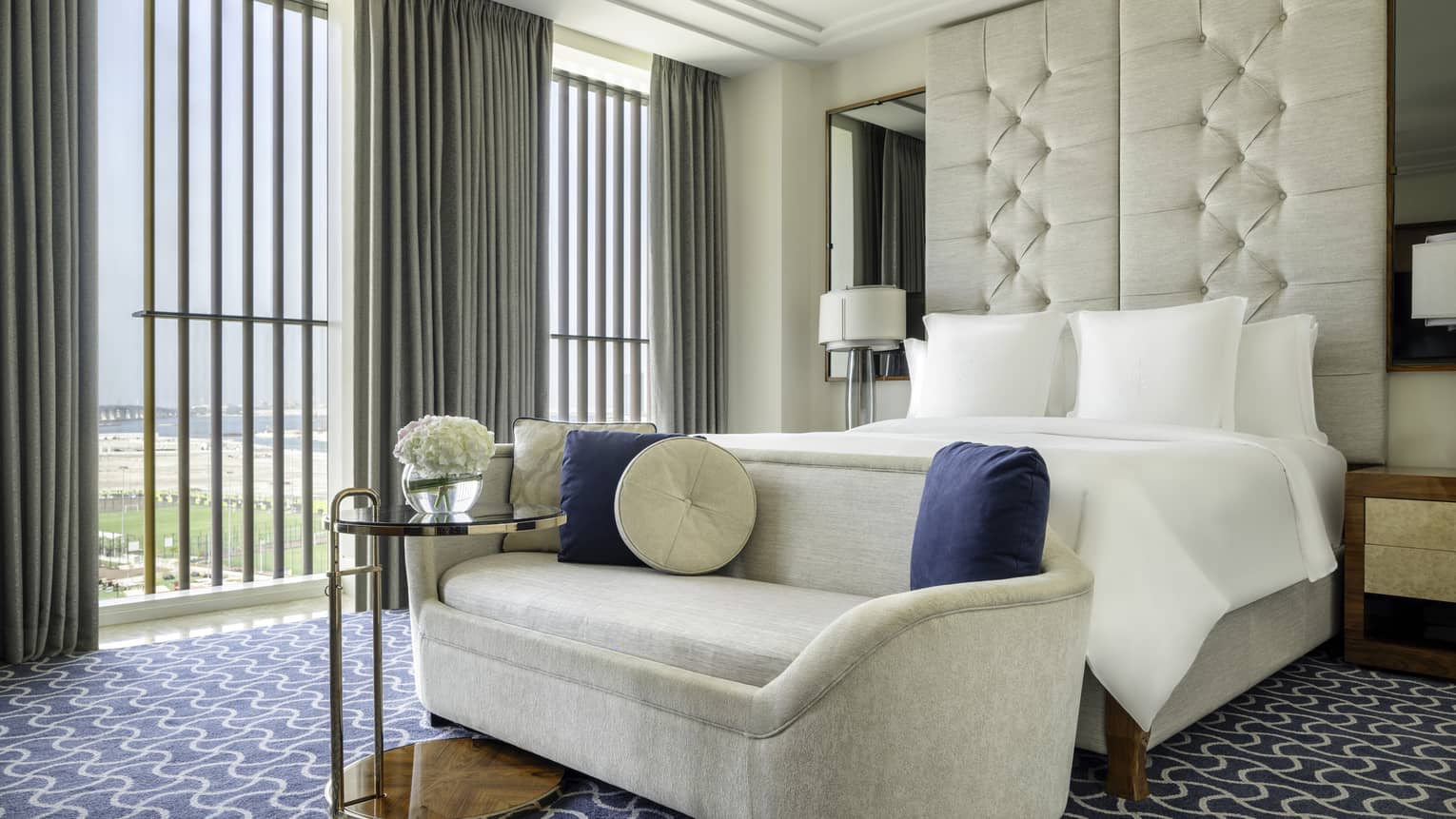 Hotel room with king bed, beige chaise at the foot of the bed, patterned carpet, ceiling-height upholstered headboard and wall of windows