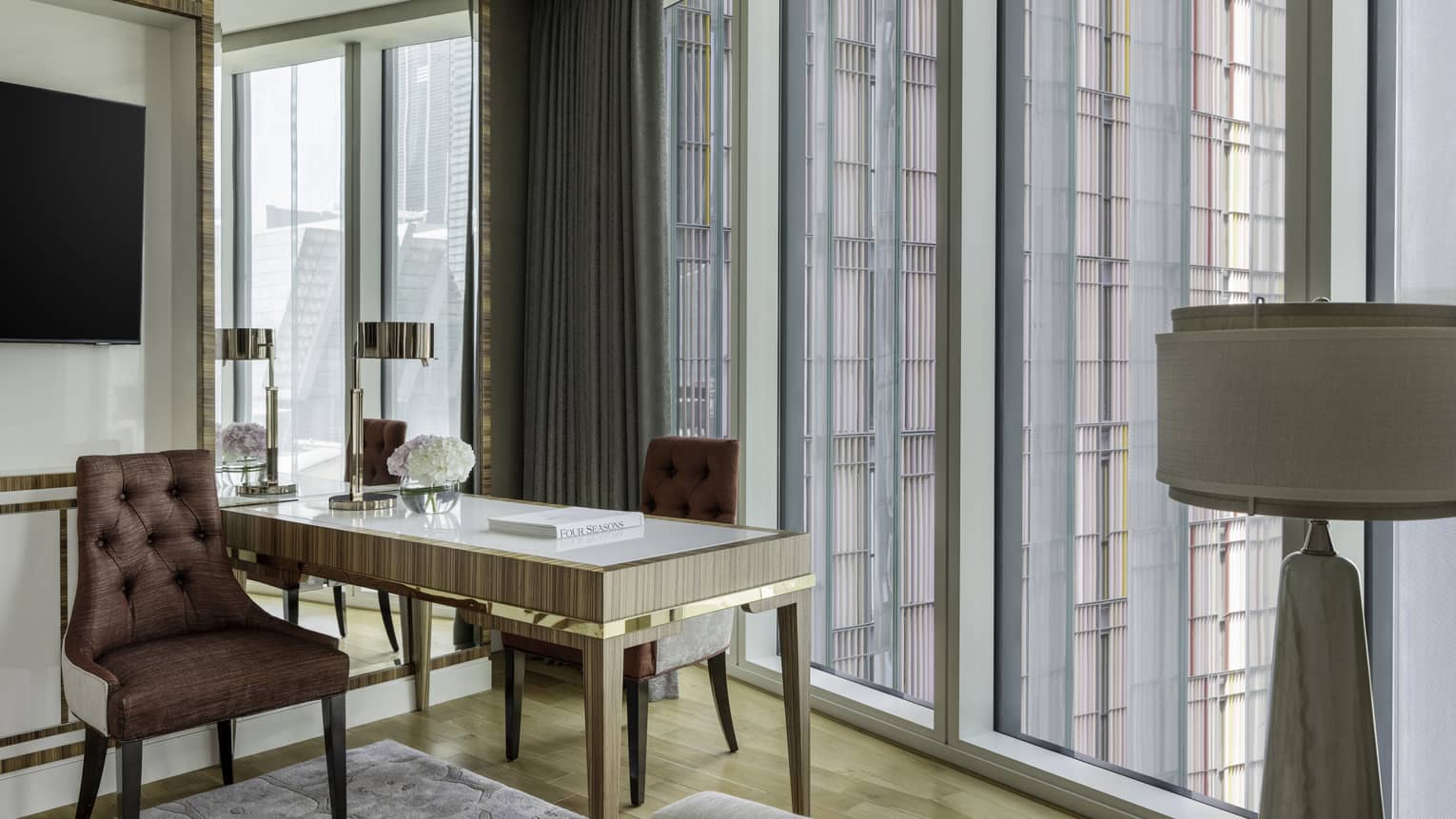 Elegant workspace with a mirrored desk, tufted brown chairs, a vase of hydrangeas and floor-to-ceiling windows offering views of a modern building facade