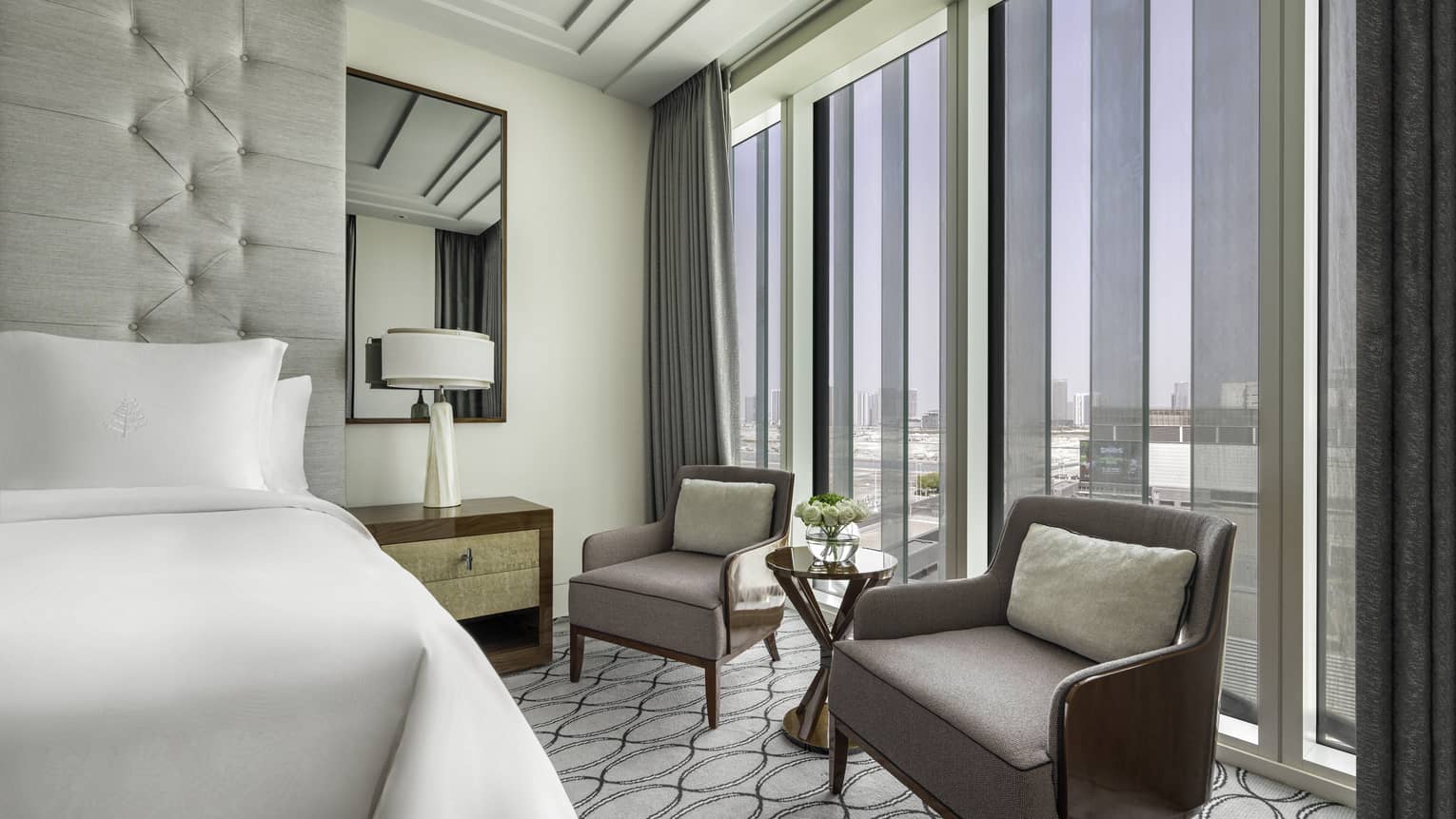 Hotel room with king bed and two arm chairs next to wall of windows