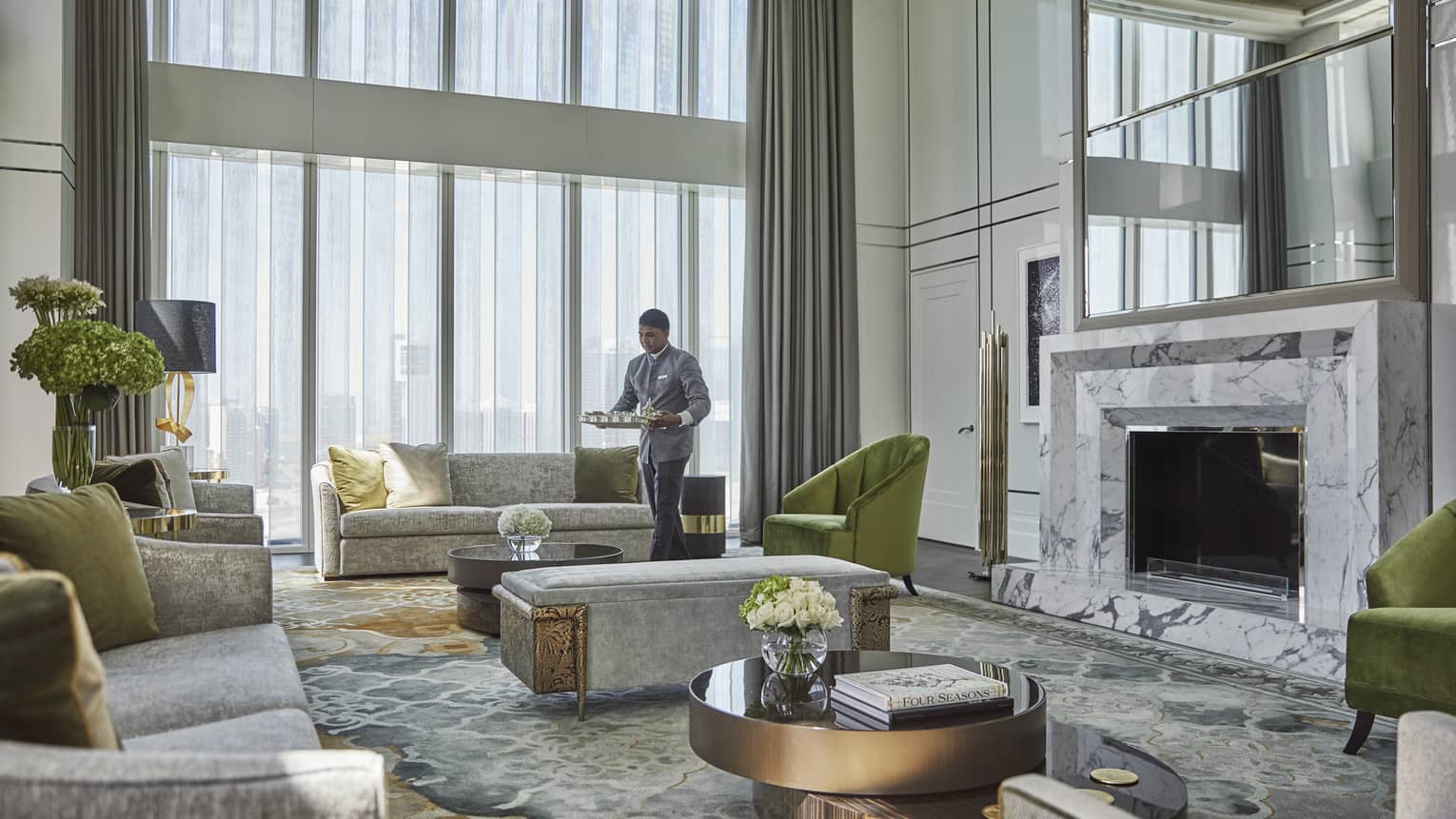 Luxurious hotel living room with high ceilings, marble fireplace, plush gray sofas, green accent chairs, floral arrangements and an attendant serving refreshments