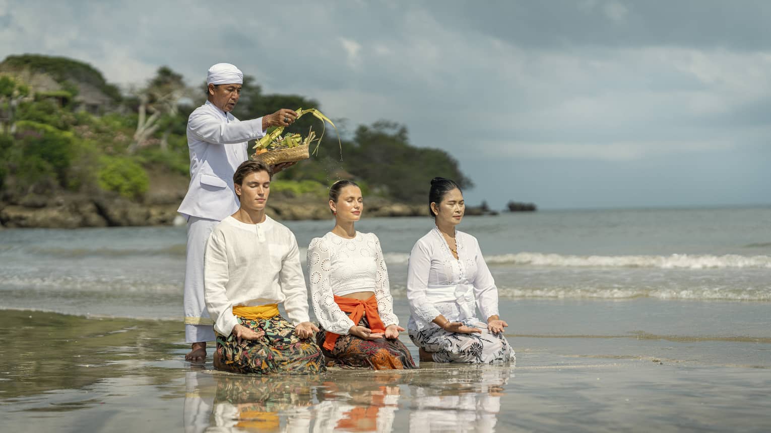 Three adults sit in shallow water with palms up on their knees as another sprinkles water from palm fronds on their heads.
