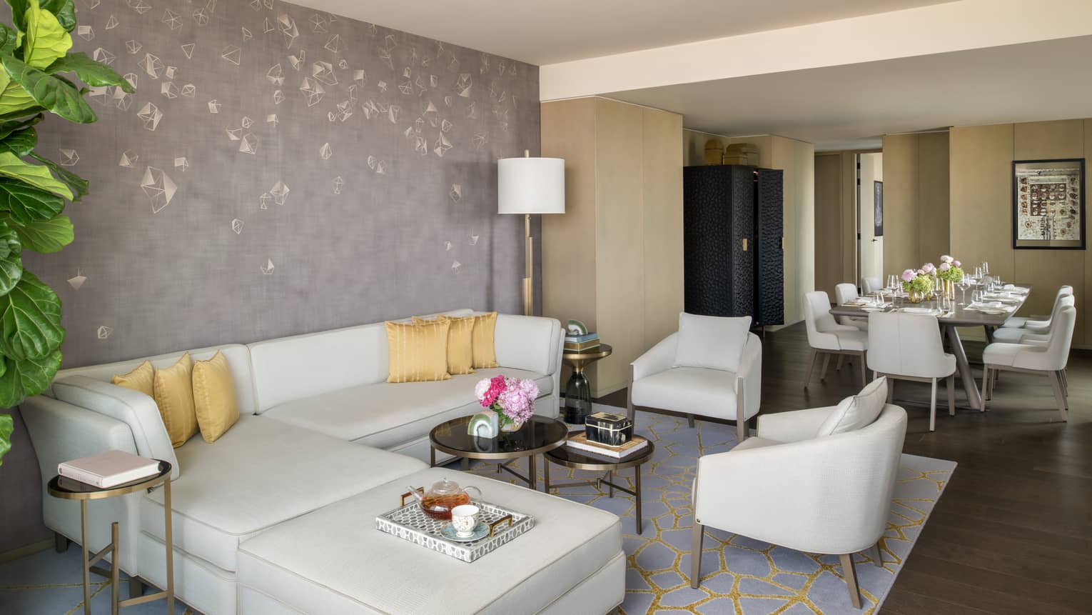 Guest room living area with cream-coloured sectional sofa and matching arm chairs, gold patterned rug, rectangular dining table in background
