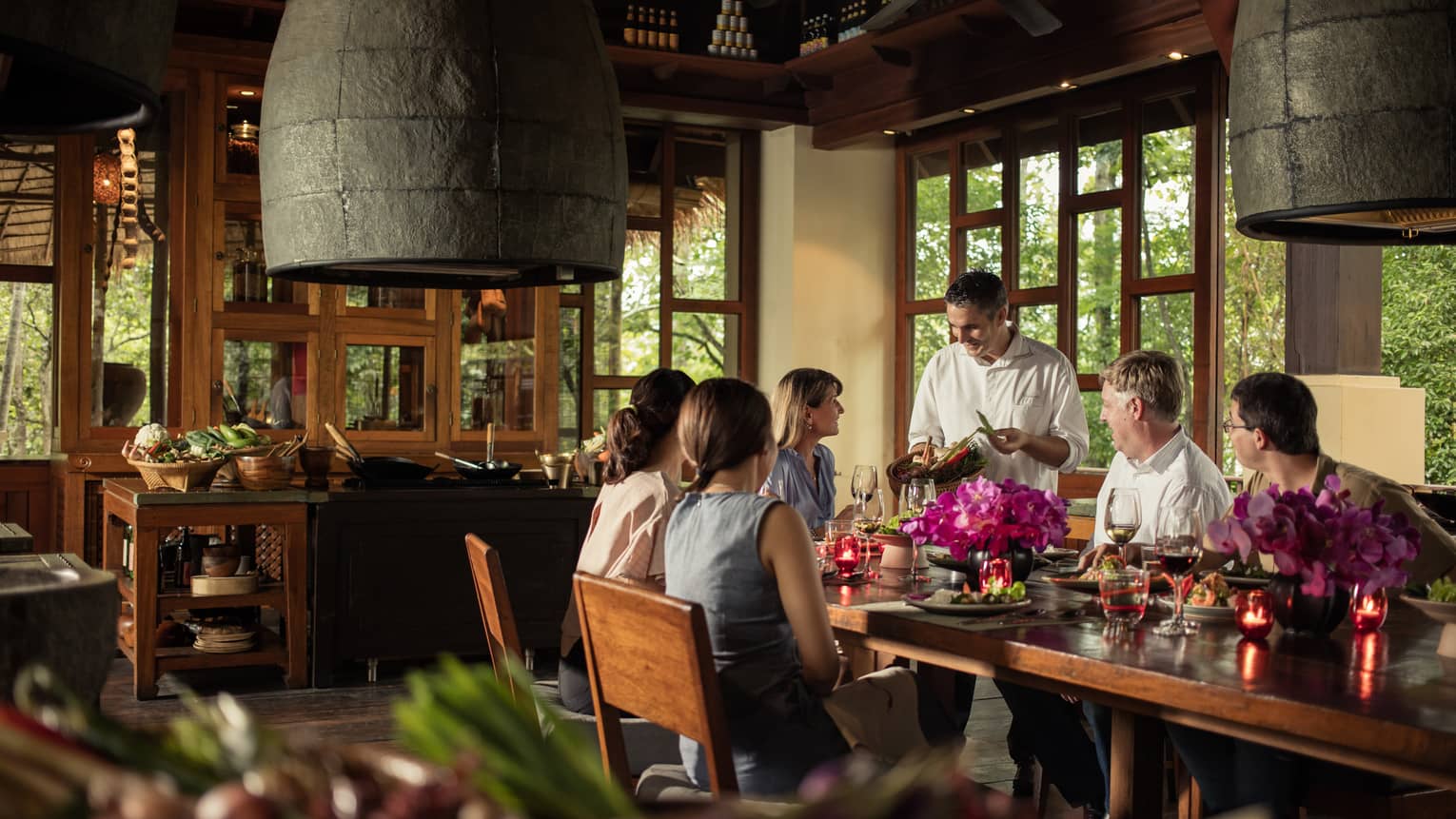 Group dining indoors with a chef presenting ingredients, surrounded by flowers, candles and tropical wooden decor.