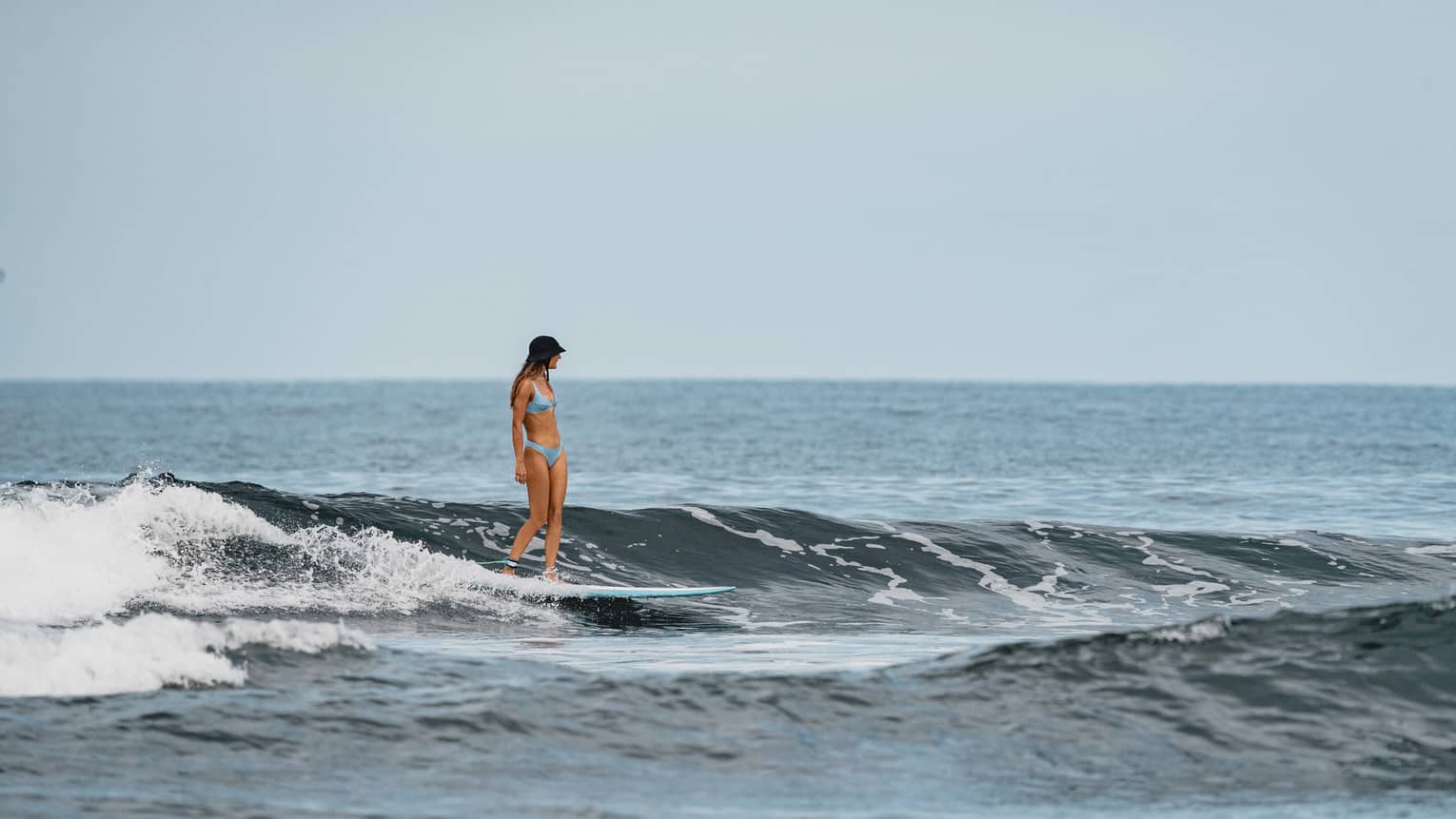 Person with long dark hair and wearing light-blue bikini rides a surfboard through the water