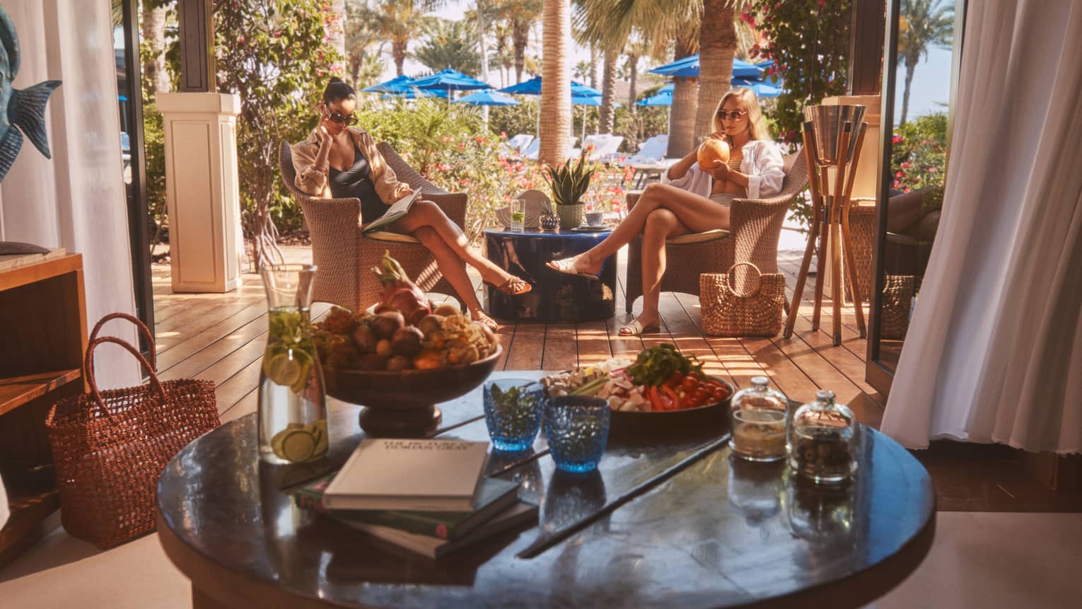 Two people sitting in rattan chairs with round table in forefront and blue pool umbrellas in backdrop