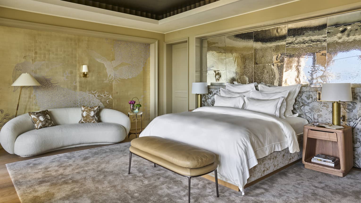 Large hotel suite bedroom featuring king bed with white linens, gold upholstered bench, side tables and lamps, white curved sofa and a silver mirrored accent wall,Large hotel suite bedroom featuring king bed with white linens, gold upholstered bench, side tables and lamps, white curved sofa and a silver mirrored accent wall