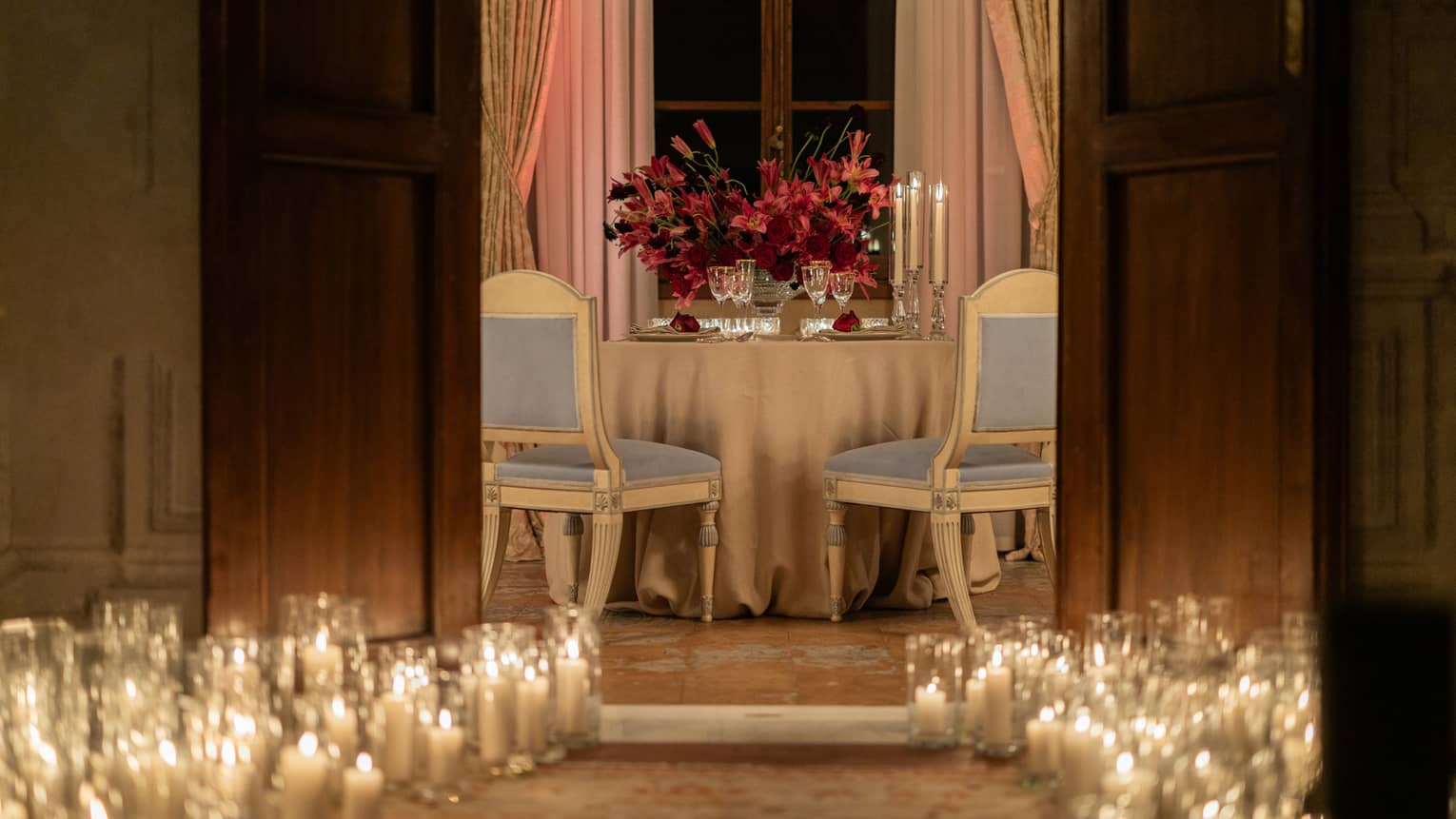 Lit hurricanes lining an entrance to a private meeting space, where a round table set for two toped with a large flower arrangement awaits