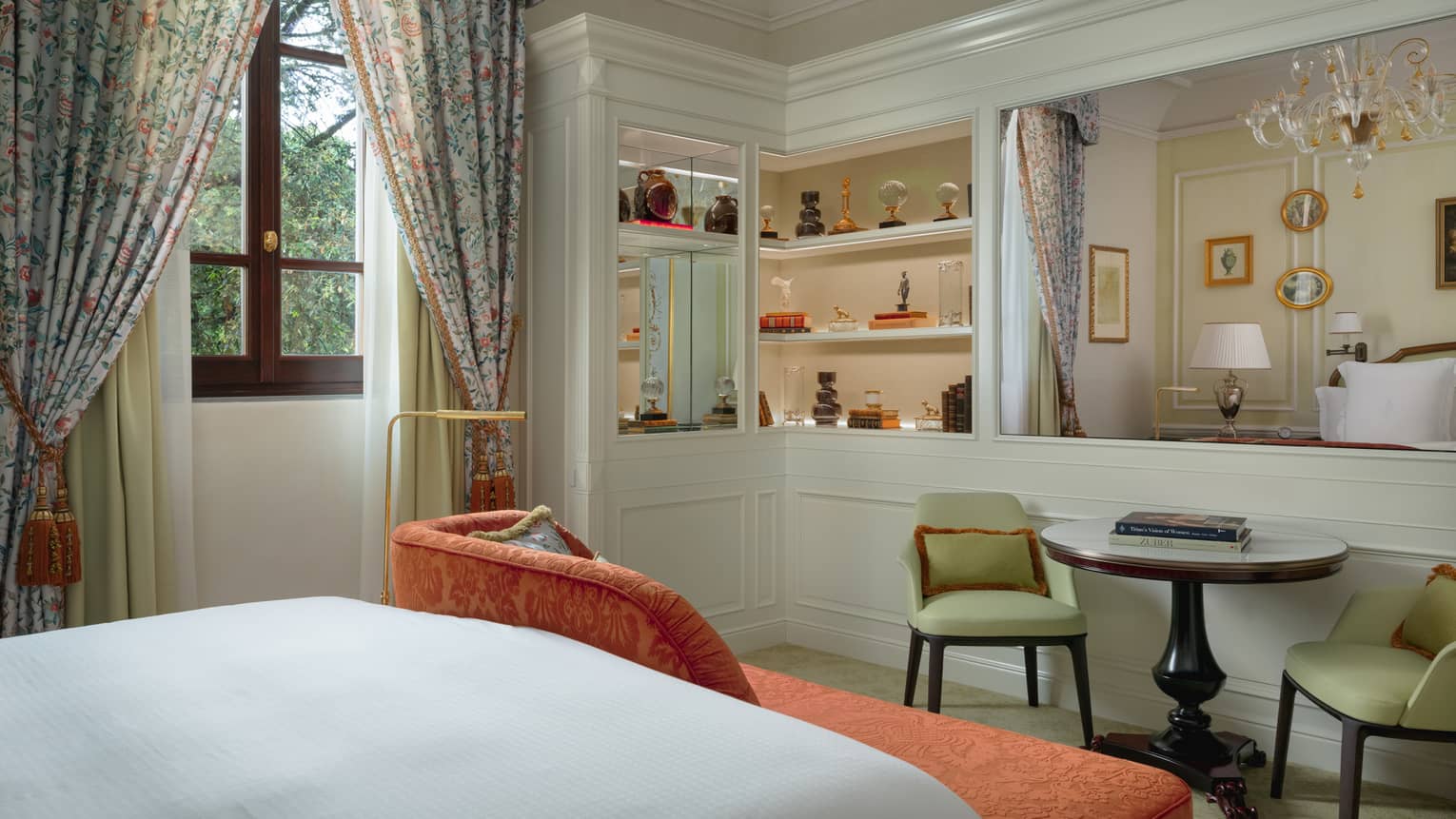 Elegant hotel room featuring a king bed, orange chaise at the foot of the bed, a table and two light green chairs, and a window with patterned curtains