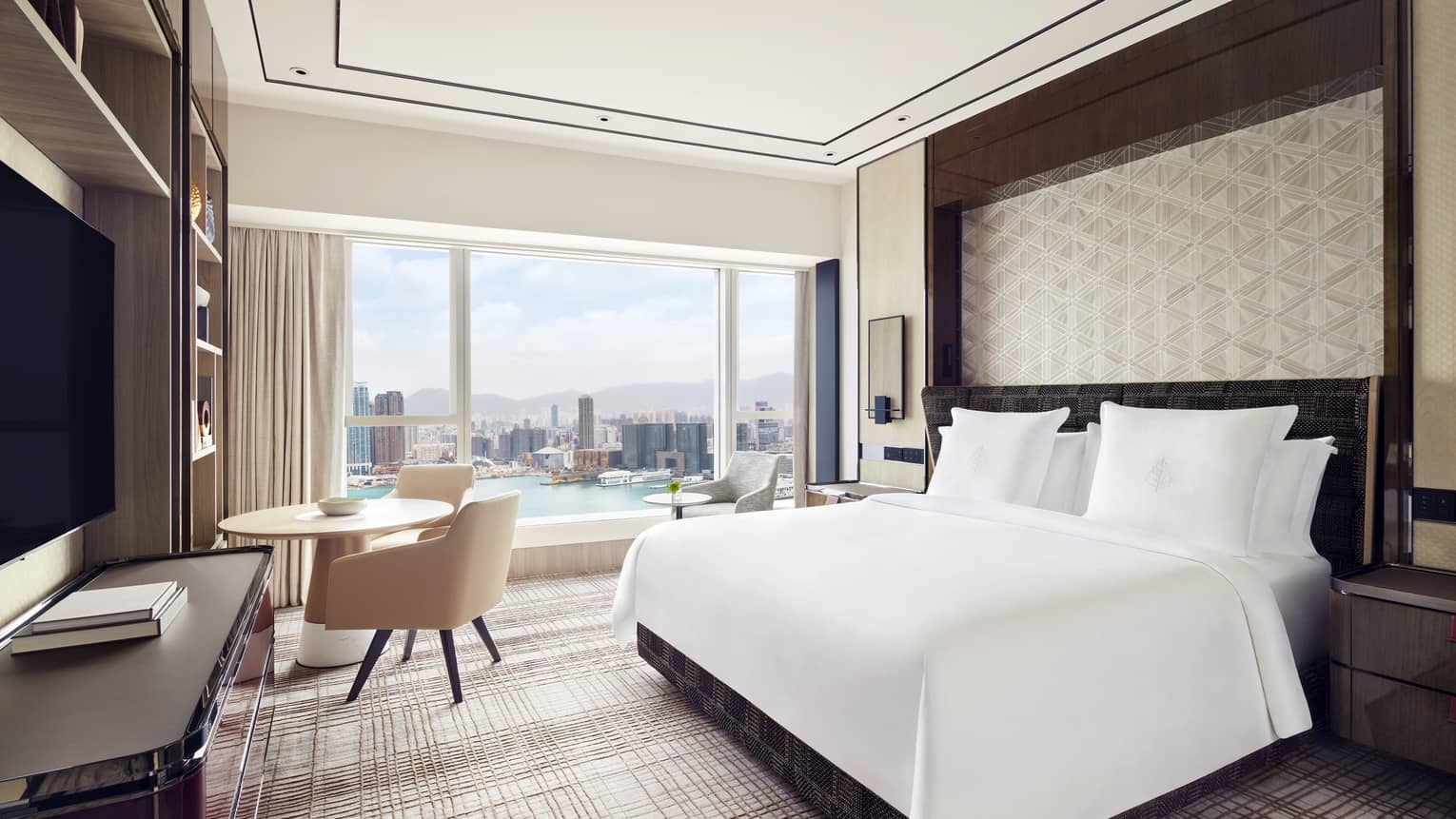 Modern hotel room with a large bed, and a round table by the window. Floor-to-ceiling windows offer a daytime view of the city skyline and waterfront.