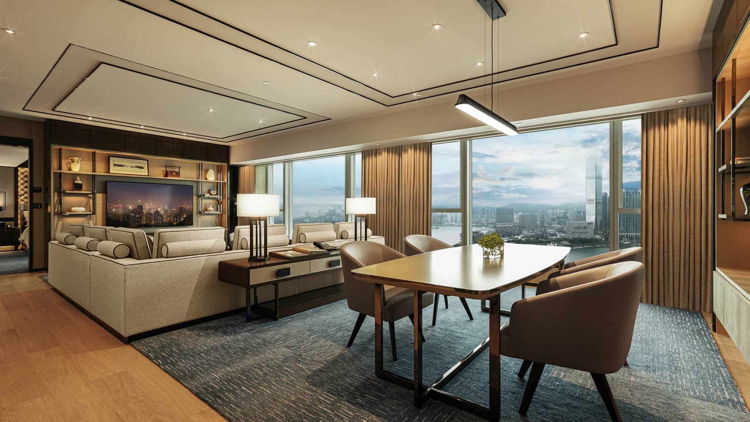Spacious hotel living area with large sectional sofa, TV, dining table, lamps, and floor-to-ceiling windows overlooking city and harbour views.