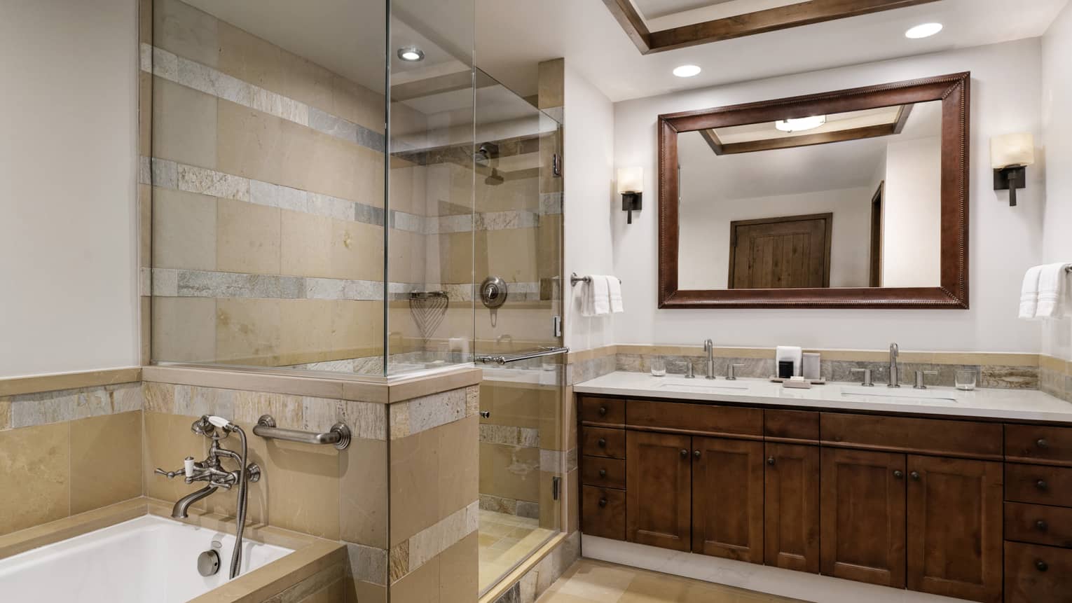 Luxury bathroom with a glass-enclosed shower, bathtub, and double vanity with a framed mirror and dark wood accents