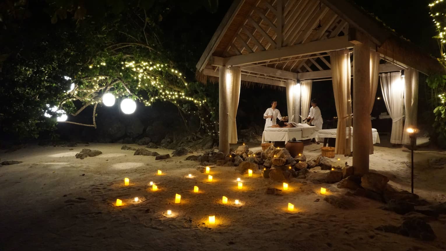 Spa treatment taking place under pergola in outdoor setting at night, with illuminated candles arranged on the ground