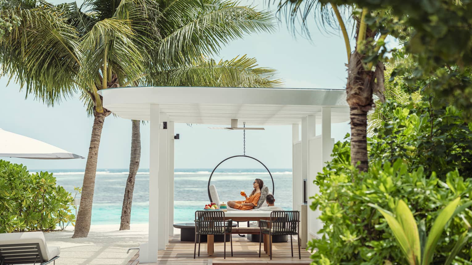 A person lounges under pergola on pool deck of private villa next to beach