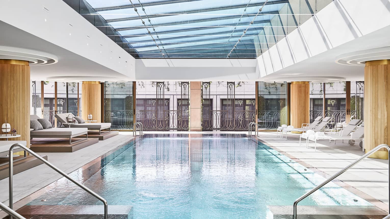 Luxury hotel indoor pool area with glass ceiling, metal railing accents and lounge chairs, surrounded by large windows and architectural detailing