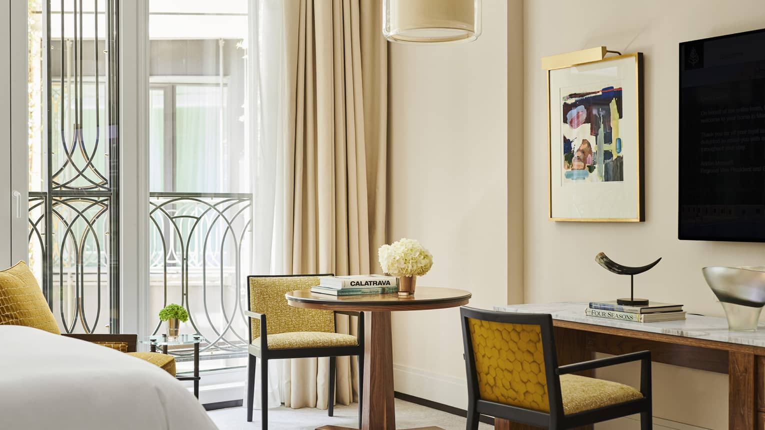 Hotel room seating area with round table, books, flowers, yellow chairs, abstract wall art, marble desk with horn sculpture and large windows with curtains