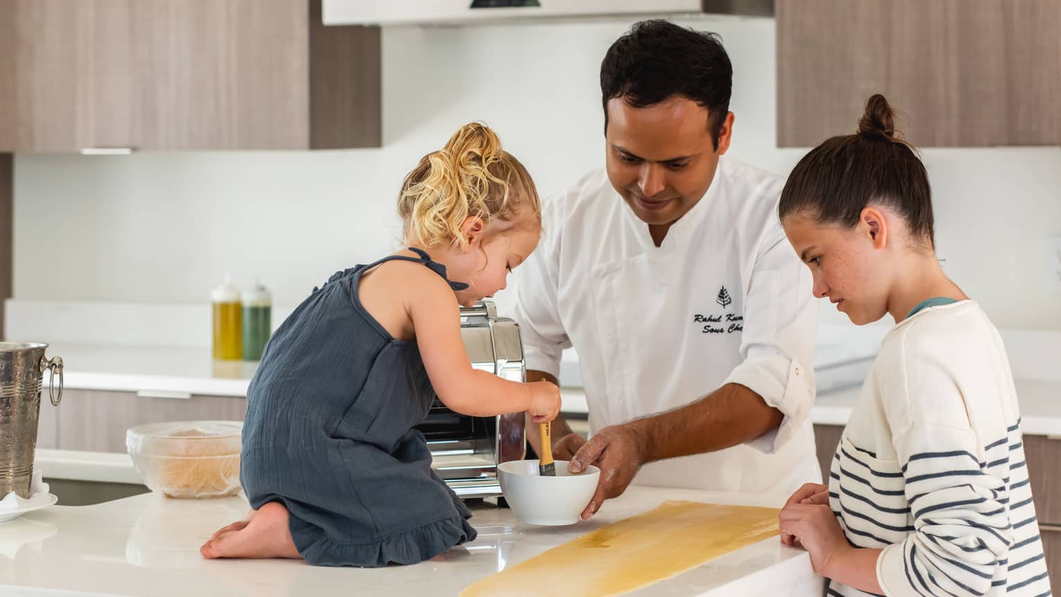 A chef helps a toddler and an older child make pasta on a white marble countertop