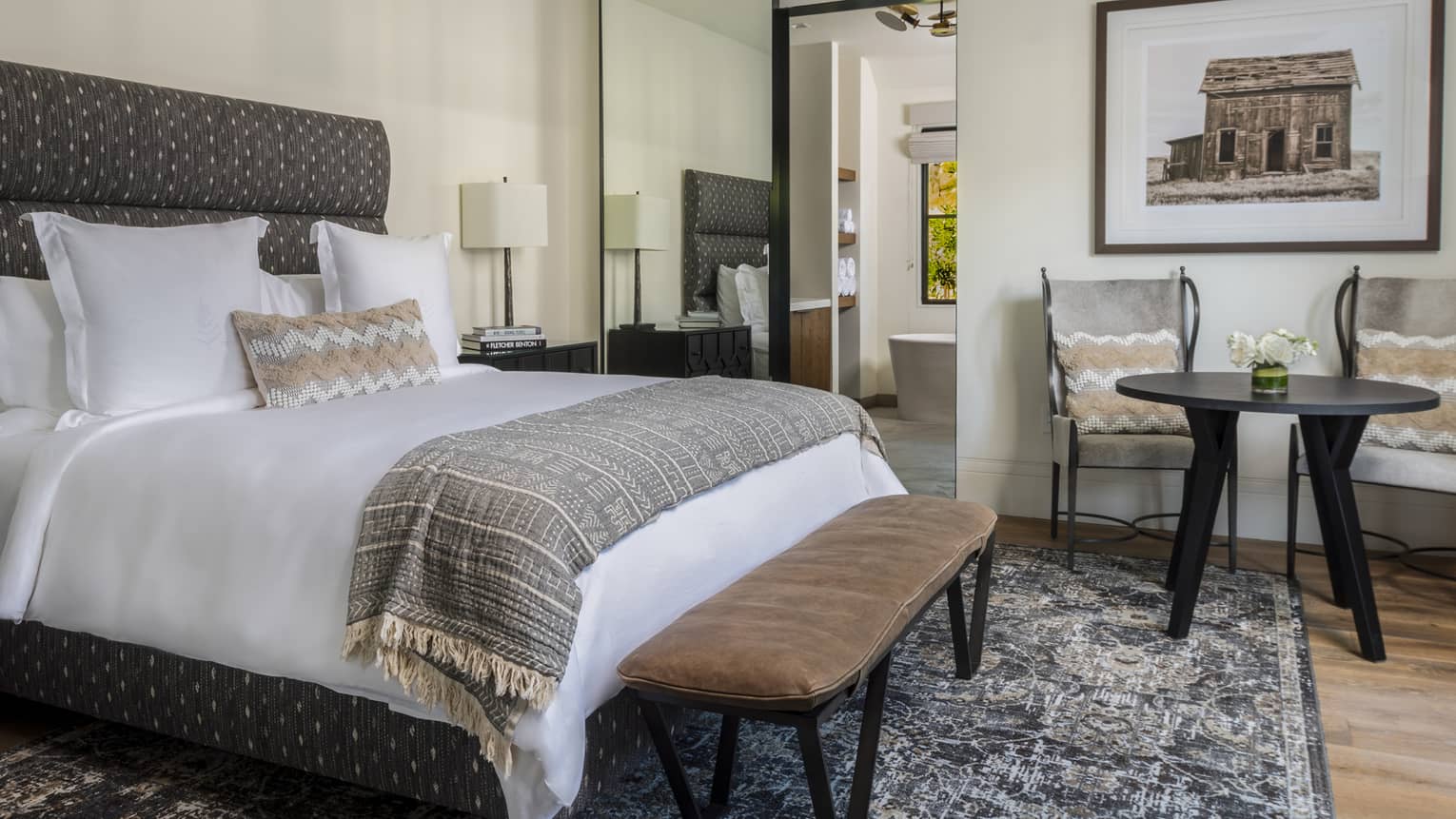 Cozy bedroom with bed with upholstered headboard, patterned throw, bench, two chairs with table, rustic wall art and view into a bathroom with freestanding tub