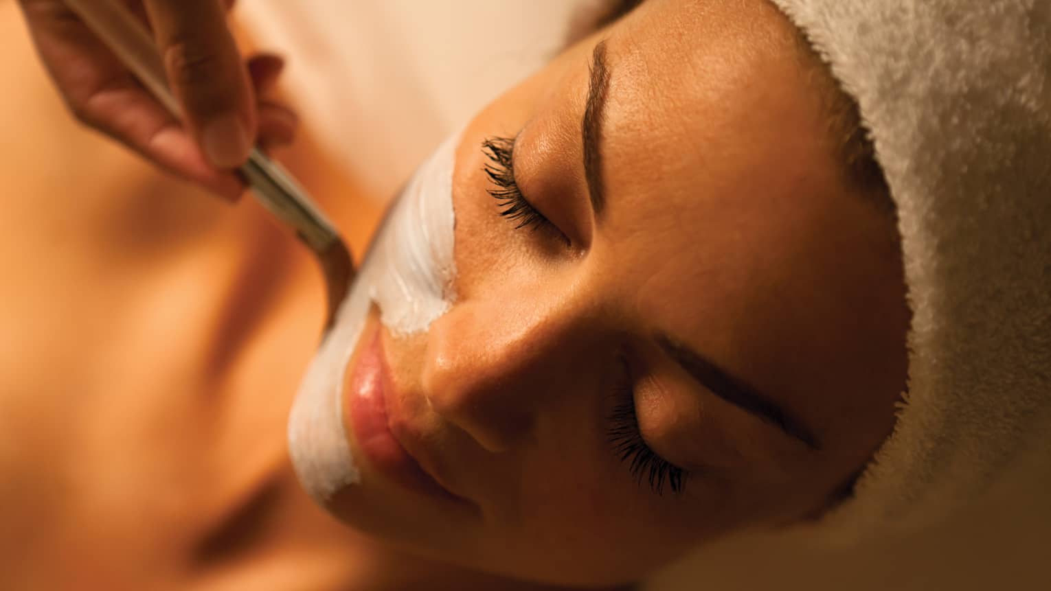 Woman with eyes closed receives a facial in dimly-lit spa room