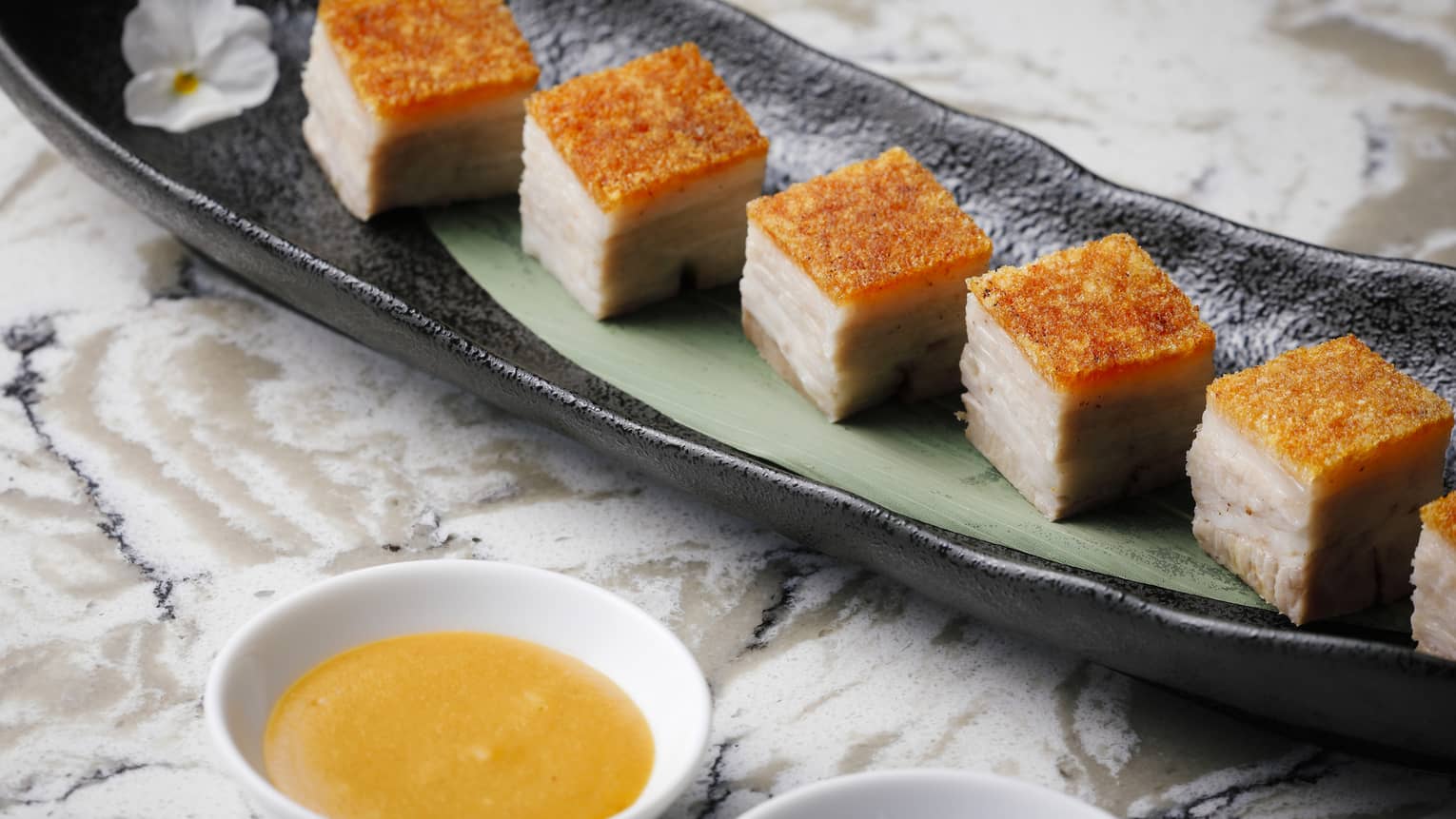 Cubes of crispy pork belly on elongated plate with dipping sauce in nearby bowl