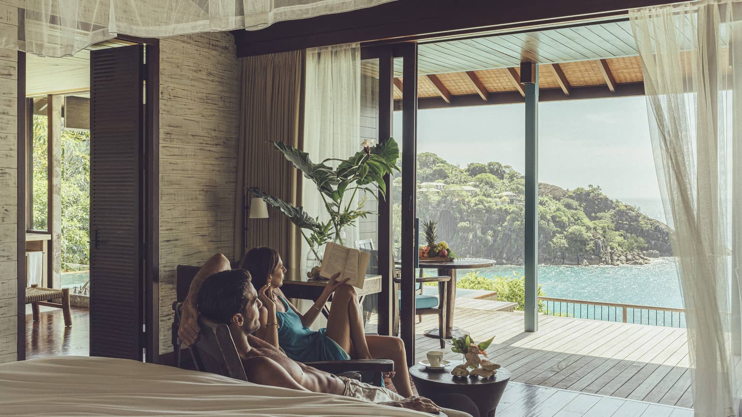 Two people relaxing in villa bedroom looking out to covered deck with small table and water views