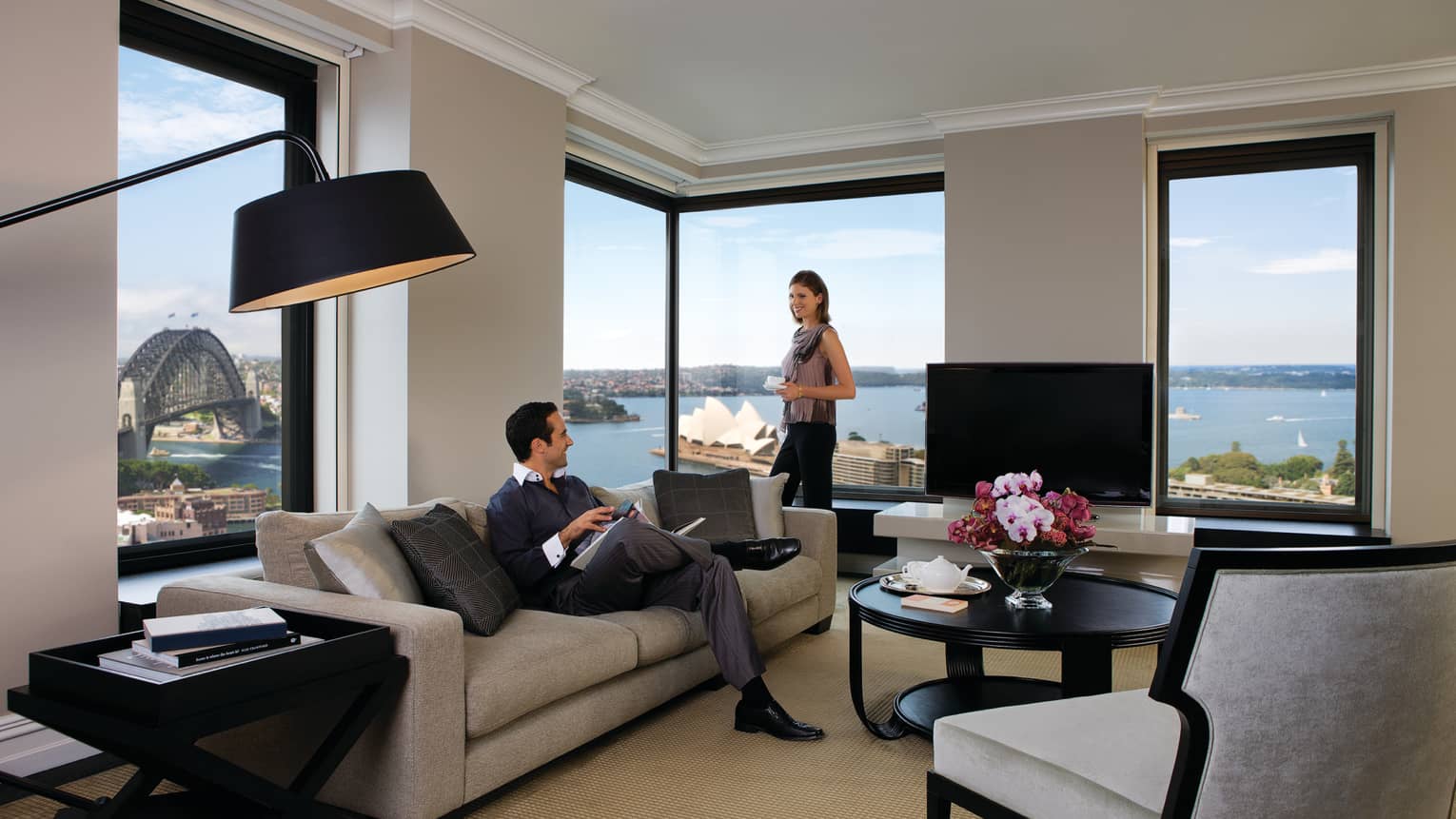 Presidential Suite, woman stands at corner windows, man reads magazine on sofa