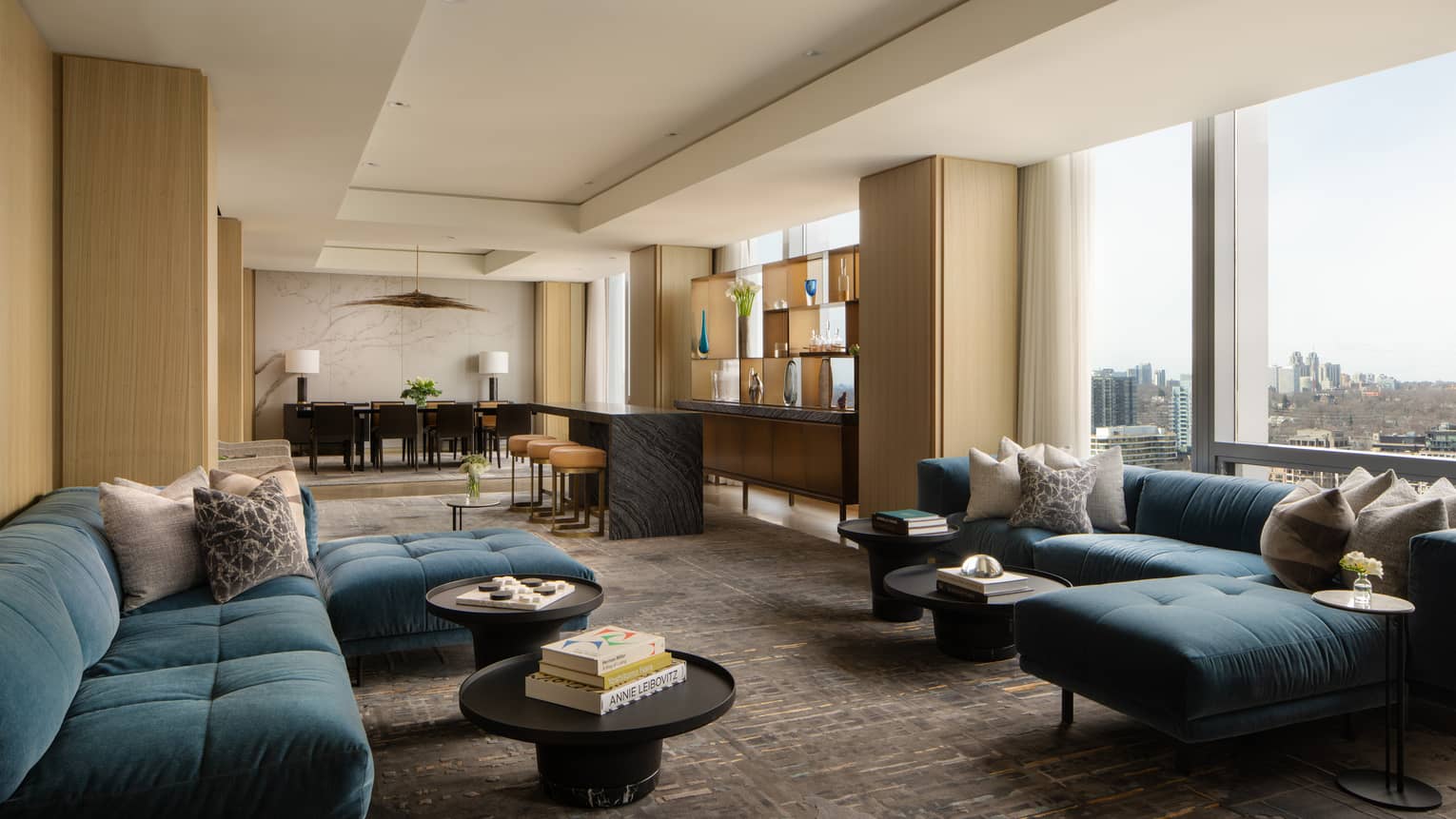 Luxury suite living area with blue sectional sofas, round coffee tables, bar with stools, dining table and floor-to-ceiling windows with city views