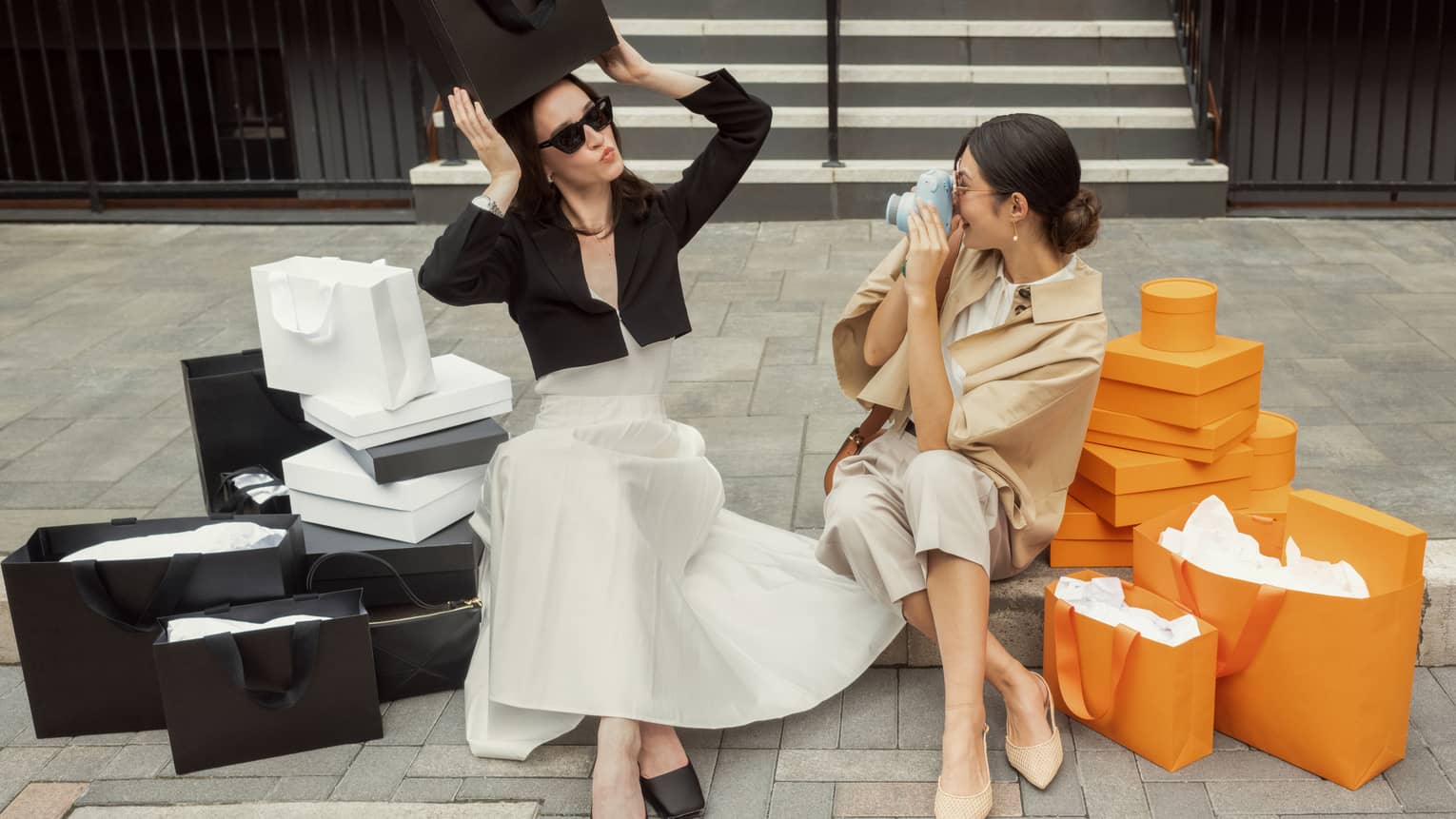 Flanked by black-and-white and orange shopping bags and boxes on stone steps, a guest poses while another takes a photo.