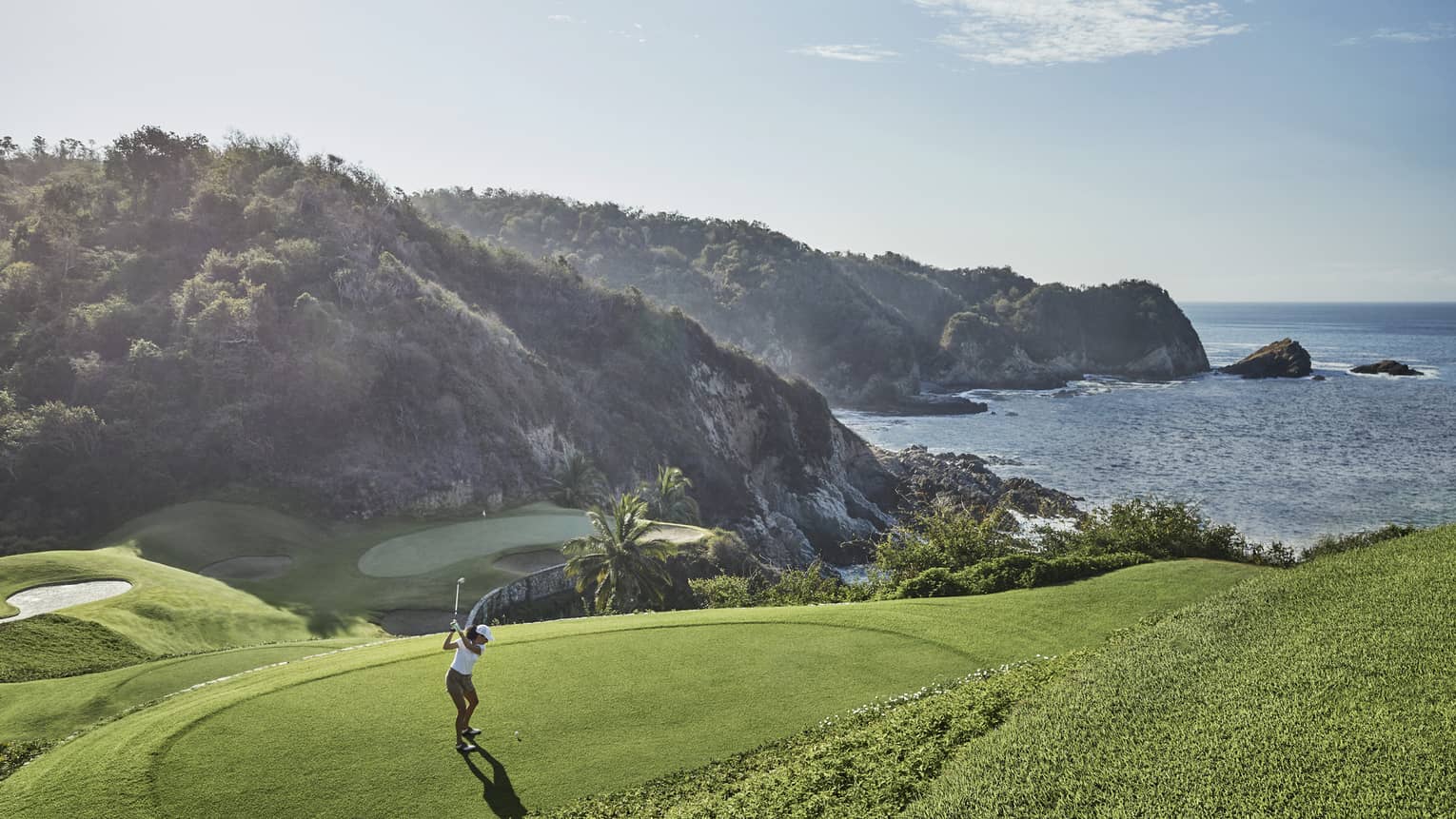 Golfer in mid-backswing, ready to strike toward a distant green at the base of an immense forested hill bordering the ocean.